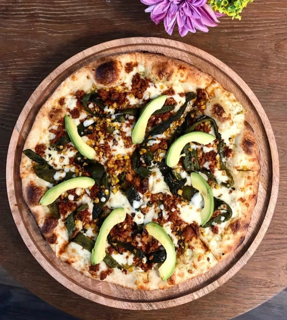 A pizza on a wooden plate with avocado and cilantro.